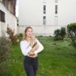 A woman stands in an urban garden holding a ginger cat on a leash, surrounded by greenery.